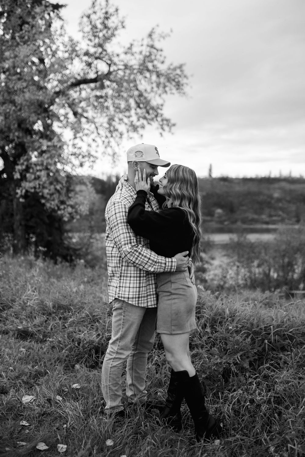 Black and white photo of an engaged couple embracing and smiling near a tree and river on a cloudy fall day.