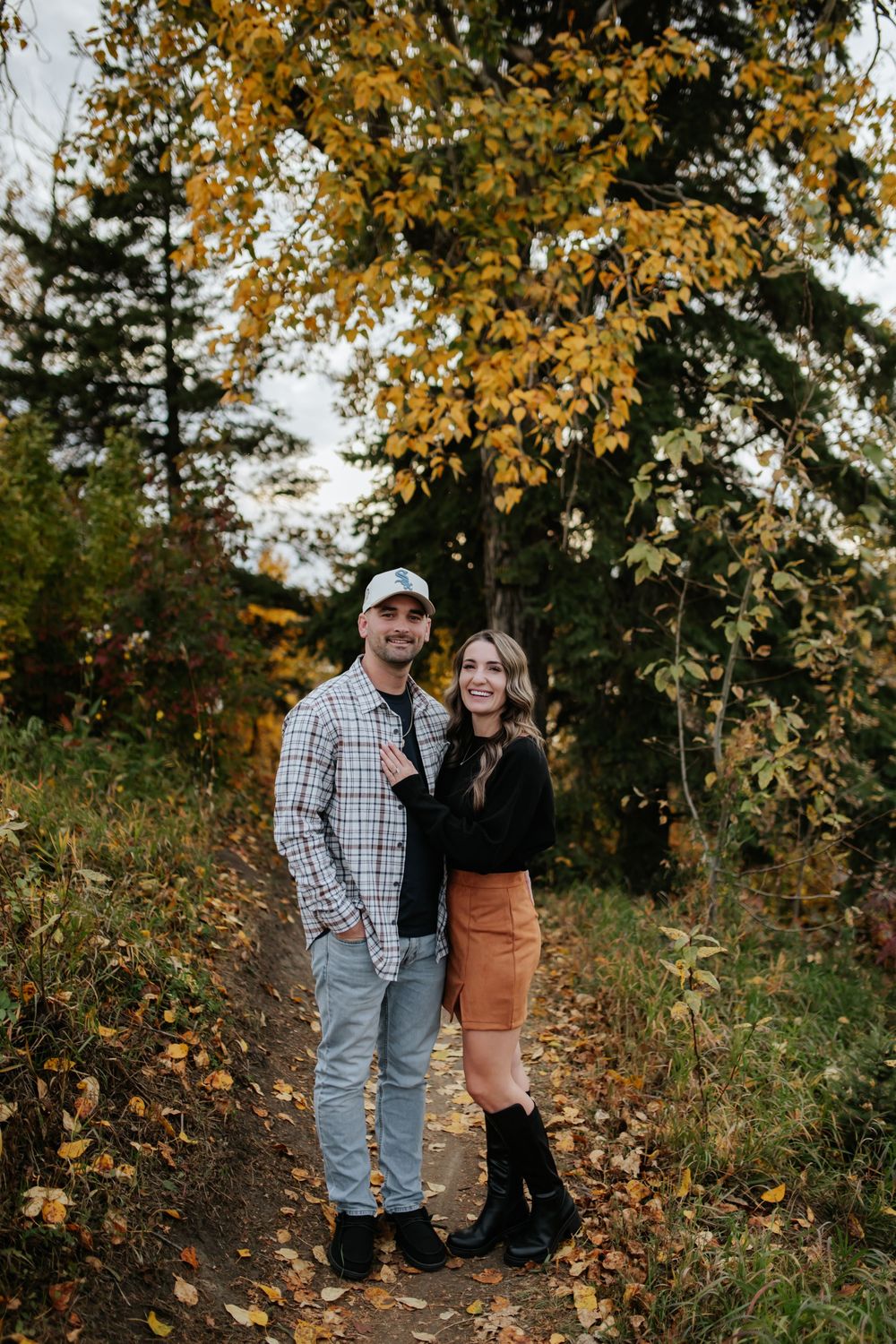Smiling couple in a plaid shirt and rust skirt embrace on a leaf-covered forest trail beneath golden autumn trees.