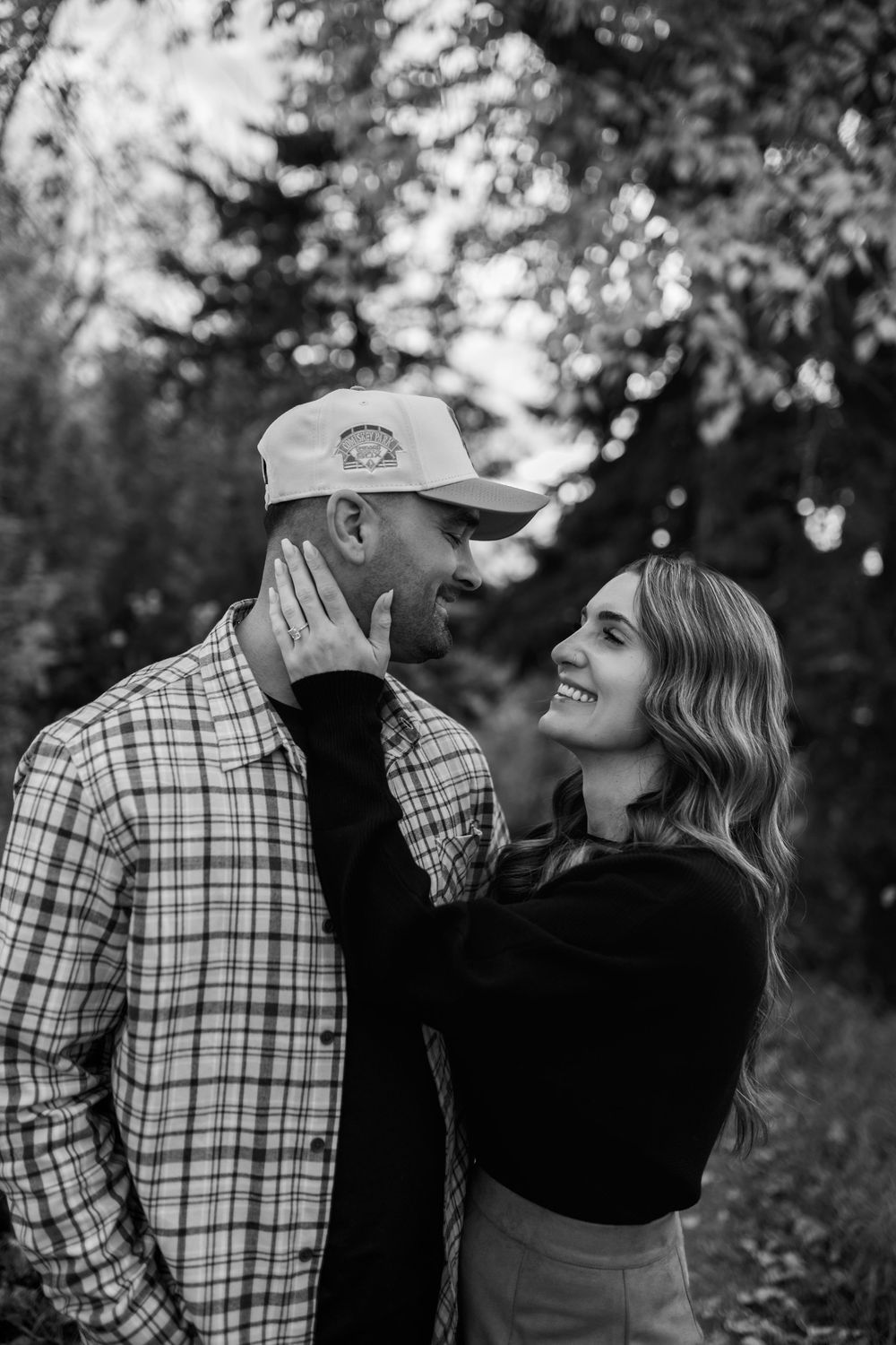 Black and white engagement portrait of a smiling couple embracing among trees, her hand on his cheek, showing off her ring.