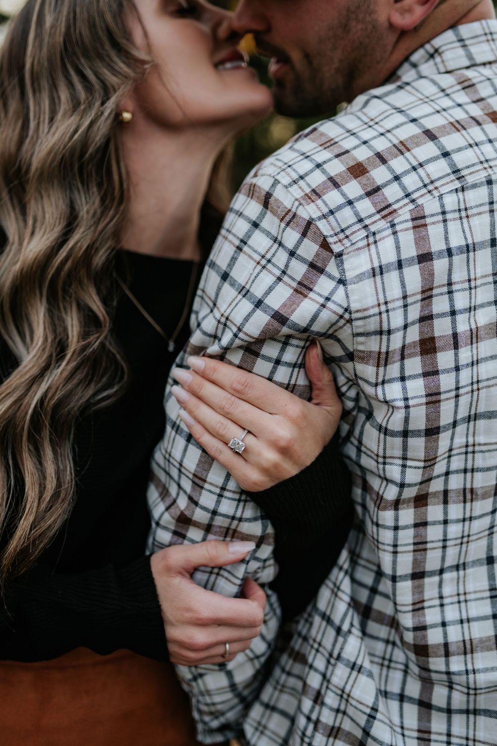Engaged couple embracing in a close-up as the woman shows her diamond engagement ring on her hand against his plaid flannel shirt.