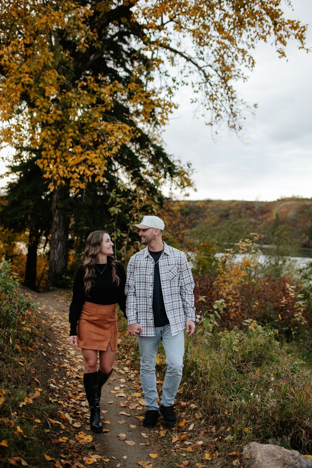 Couple in plaid shirt and rust skirt hold hands walking a leaf-covered path beneath golden autumn trees during an engagement session.