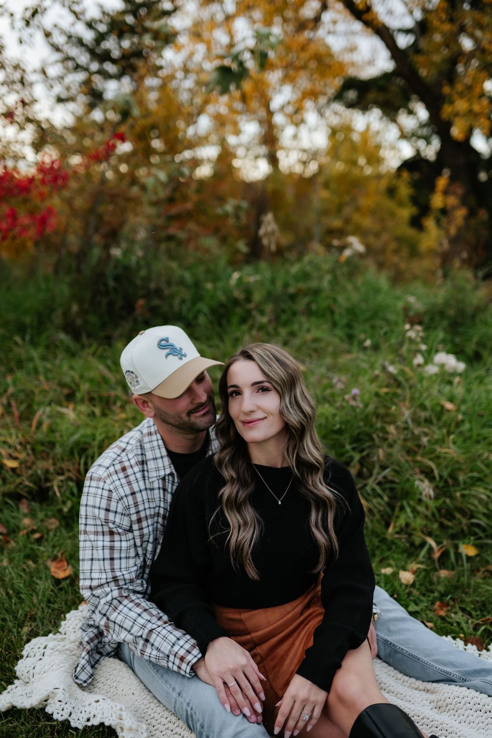 Couple cuddling on a lace blanket in a grassy field with colorful autumn foliage, the man in a cap and plaid shirt, the woman smiling.