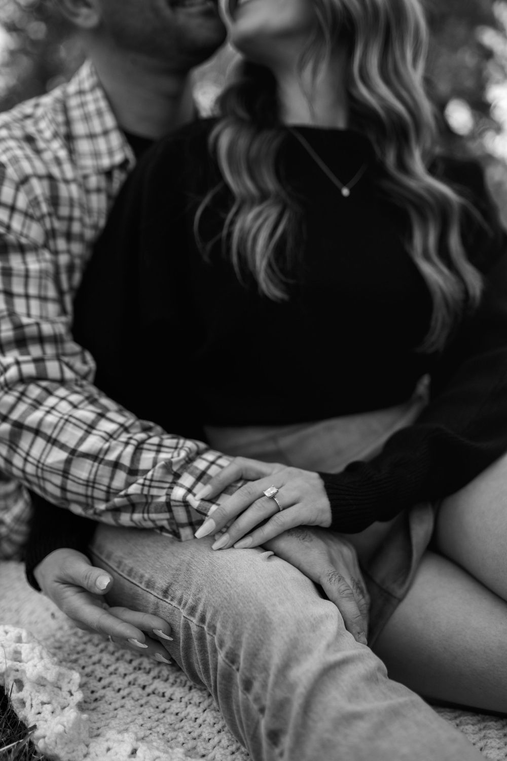 Black and white close-up of an engaged couple embracing on a blanket, her hand with a sparkling ring resting on his.