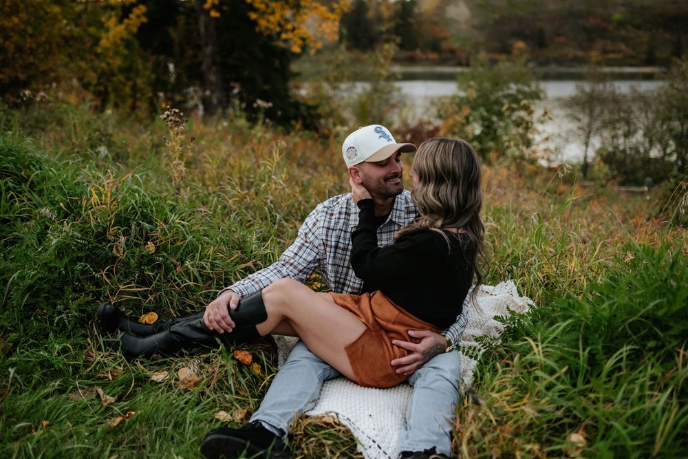 A couple in casual autumn outfits sits close together on a blanket in tall grass beside a calm lake, sharing an affectionate moment.