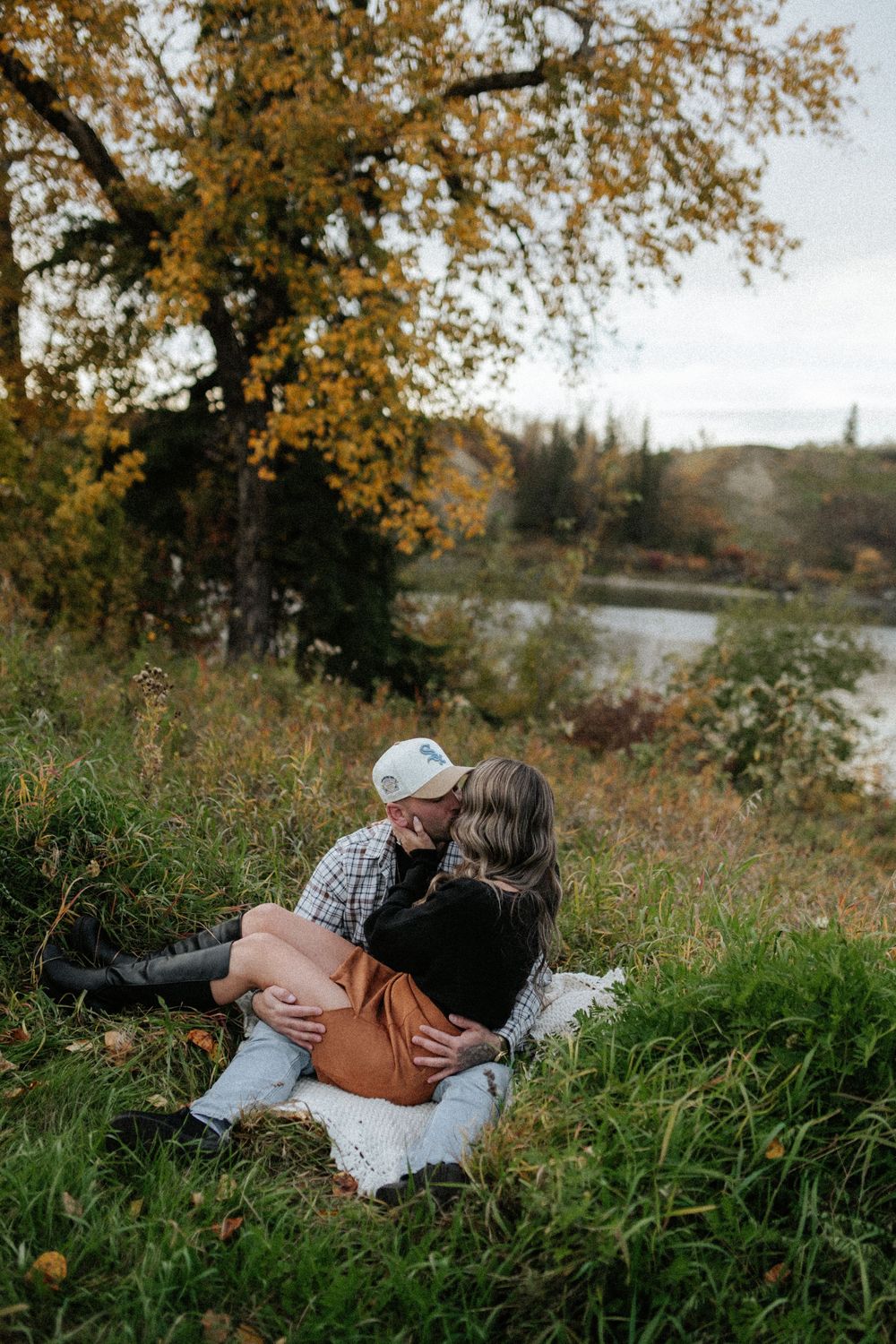 Couple embracing and kissing on a blanket in a grassy field beside a lake, with golden autumn trees in the background.