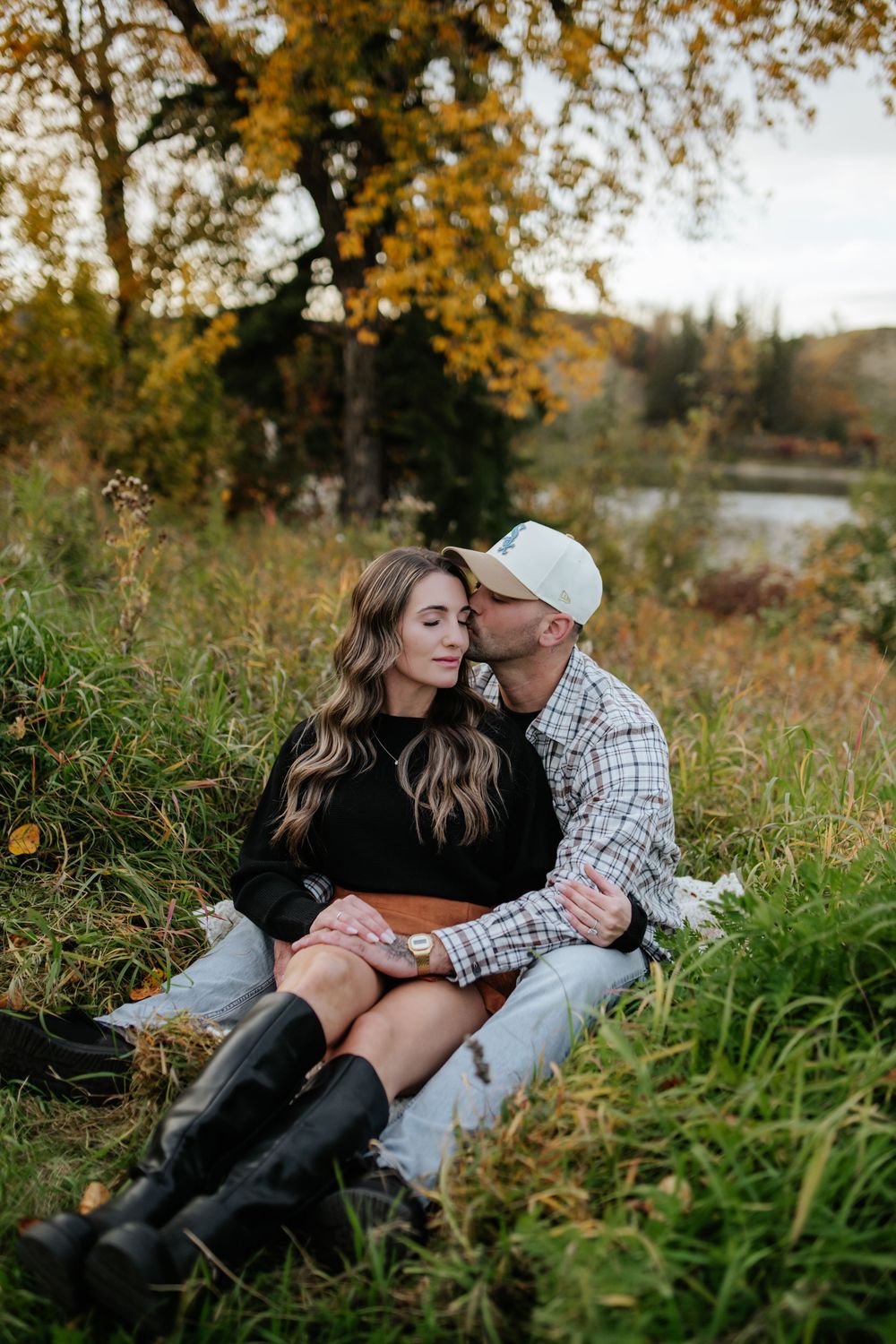 Couple sitting close in tall autumn grass as he kisses her forehead, with a lake and golden fall foliage in the background.