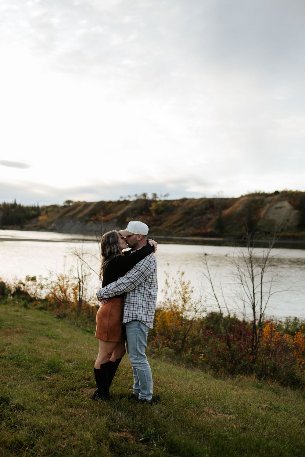 Couple in a warm embrace on a grassy riverbank, with autumn foliage and a hillside across the water under a soft overcast sky.