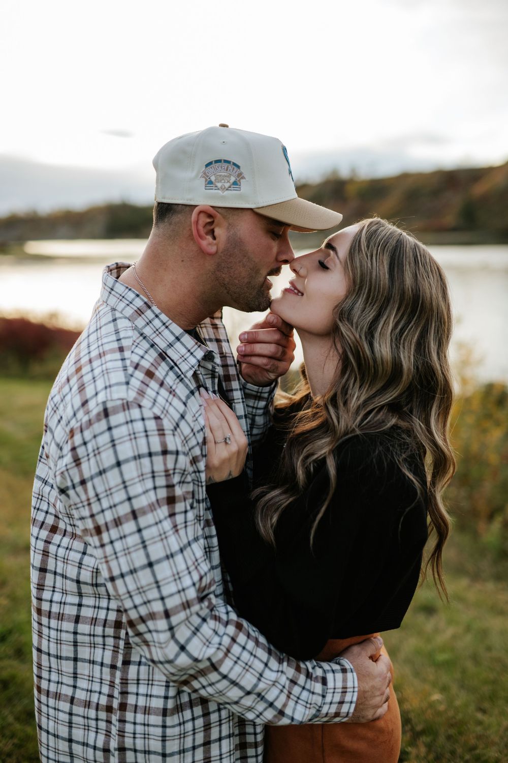 Couple in a tender embrace touching noses during an outdoor engagement photo session beside a lake at sunset.