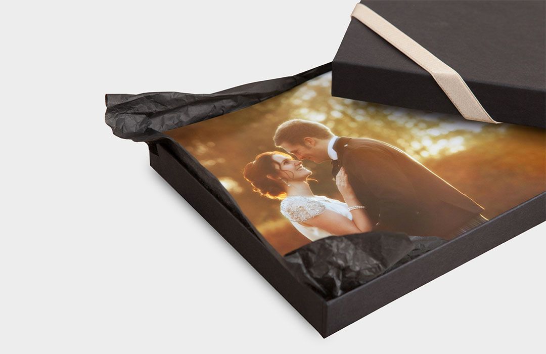 Black gift box with cream ribbon holding a framed wedding photo of a couple kissing, wrapped in black tissue paper.