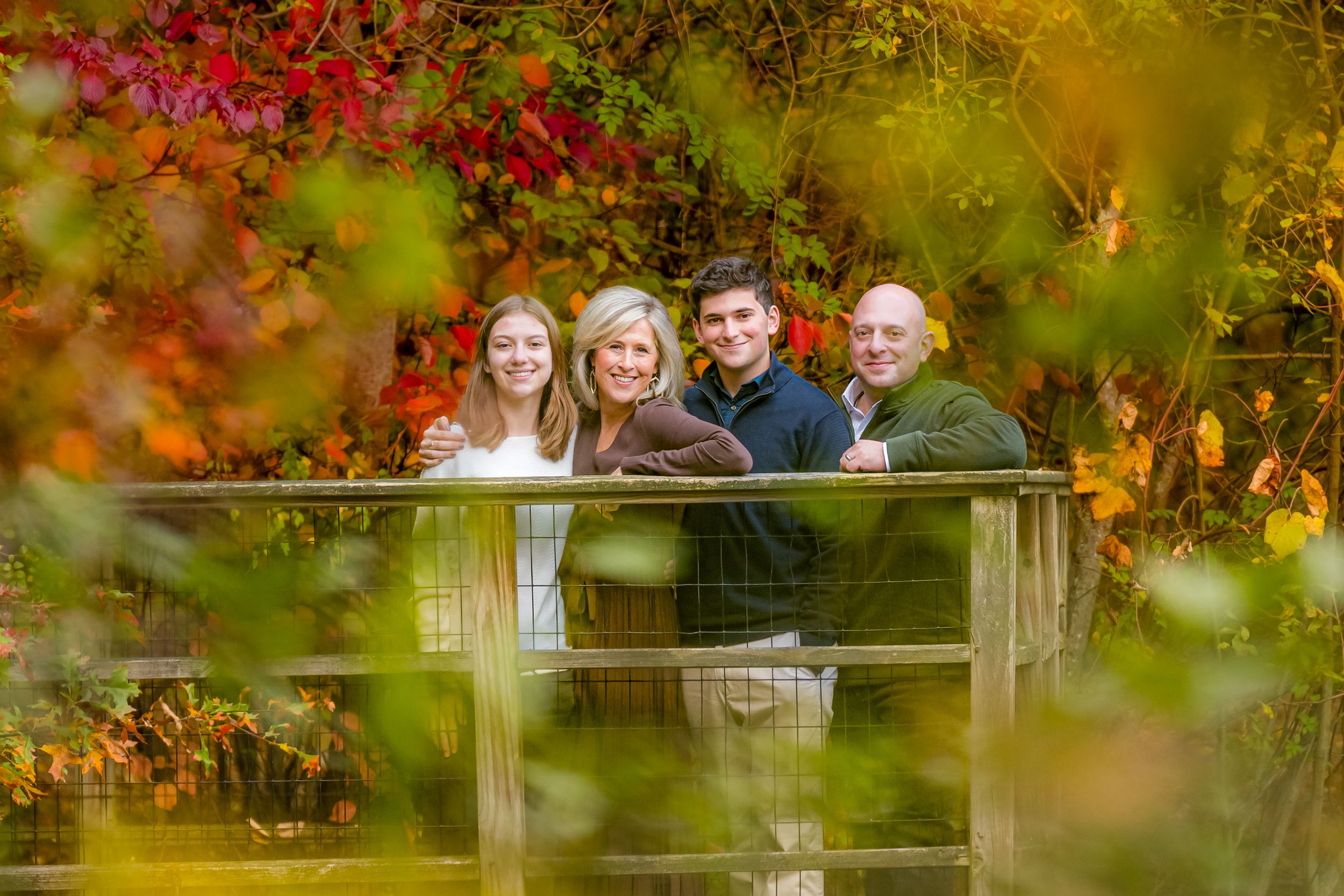 Family of four smiling together behind a wooden railing, surrounded by colorful red and orange autumn foliage.