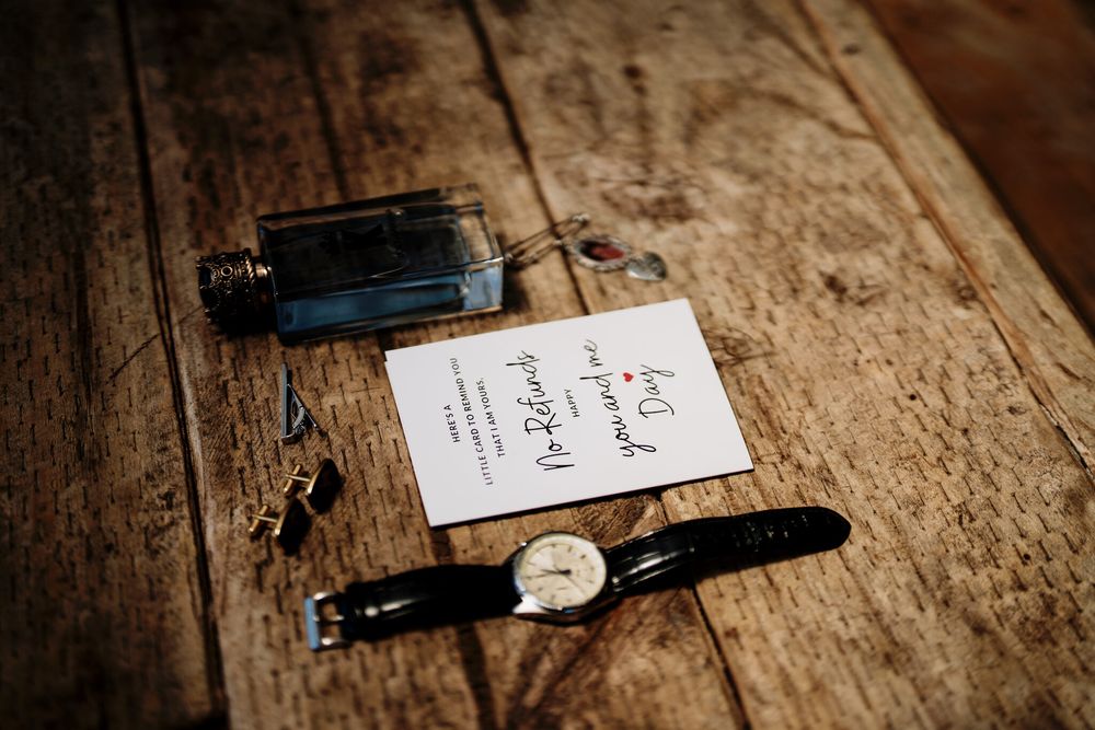 Groom's wedding day flat lay with a leather-strap watch, cufflinks, cologne bottle and a handwritten vow card on a rustic wooden table.