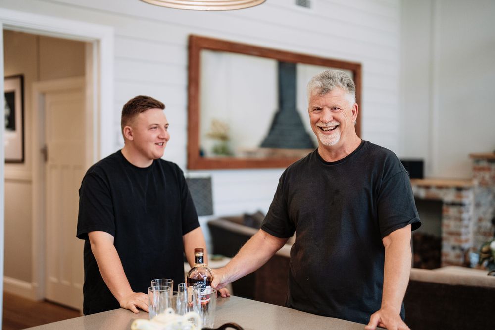 Two smiling men in black t-shirts stand at a kitchen counter with glasses and a bottle in a bright, rustic room with a fireplace.