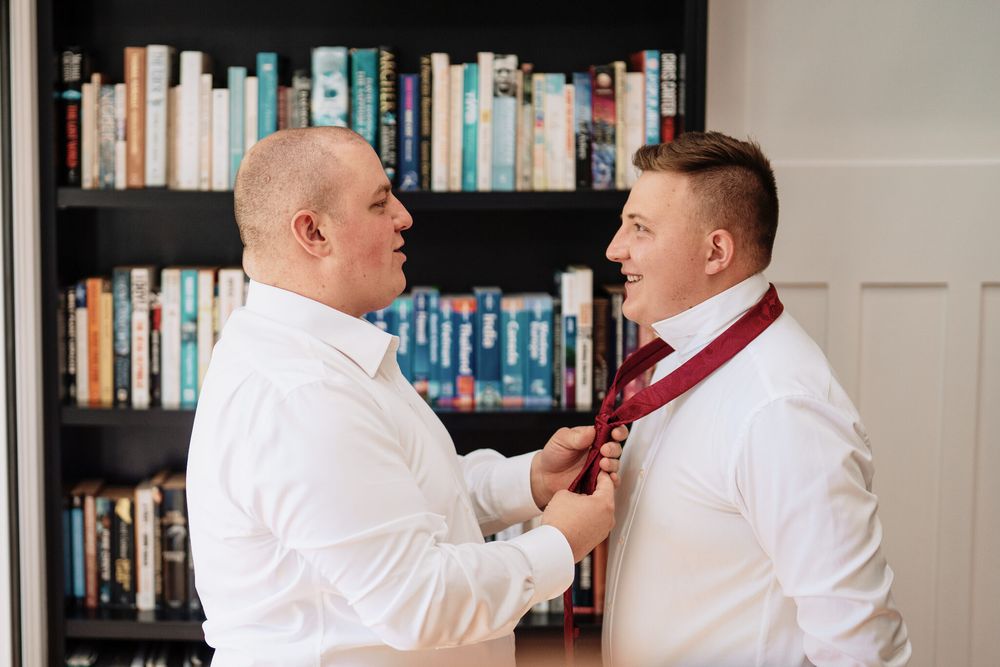 Two men in white dress shirts stand in front of a bookshelf as one helps the other adjust a burgundy tie.