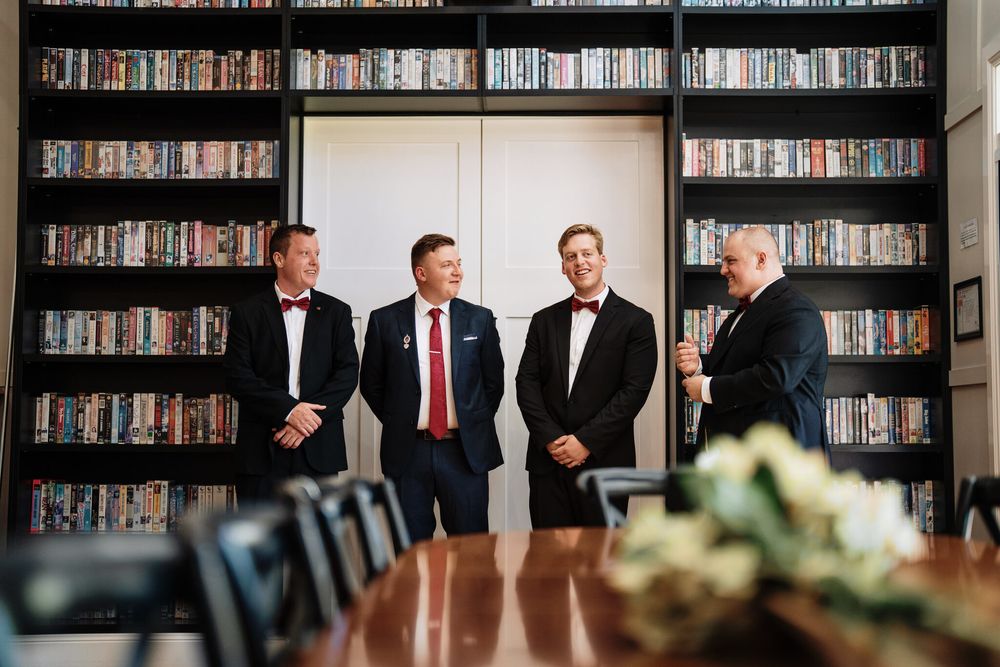 Four men in dark suits stand in a row in front of tall bookshelves, with a polished wooden table and white flowers in the foreground.