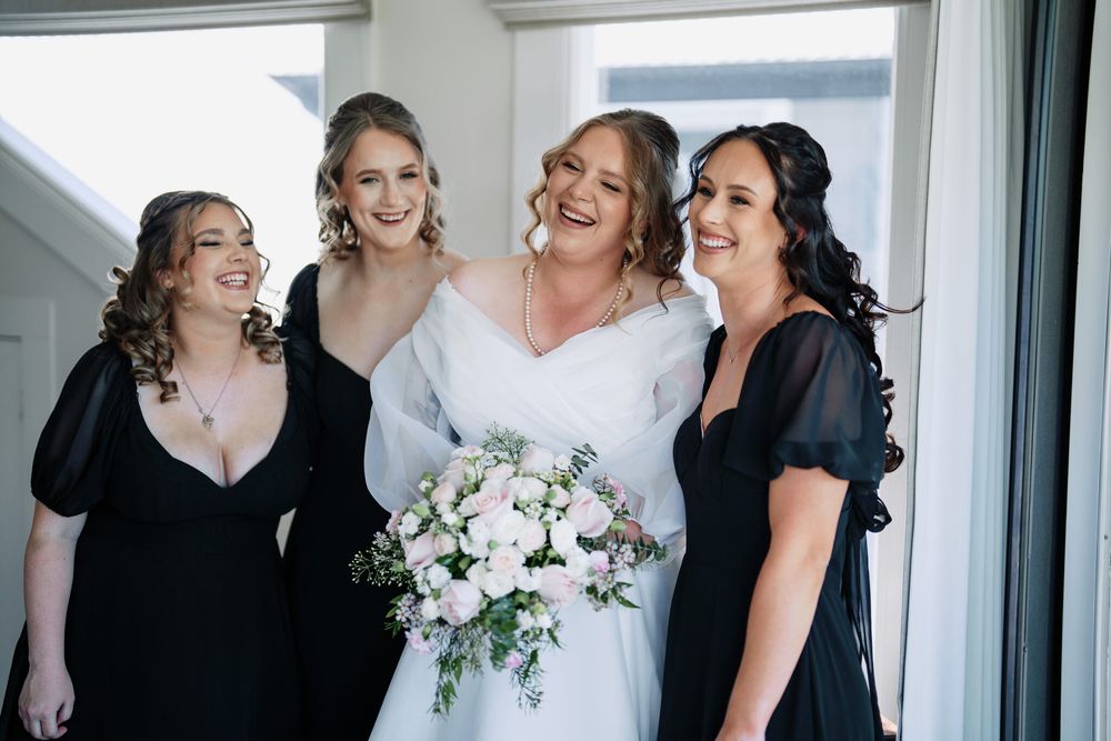 A smiling bride in a white gown holds a white rose bouquet, laughing with three bridesmaids in black dresses beside a bright window.
