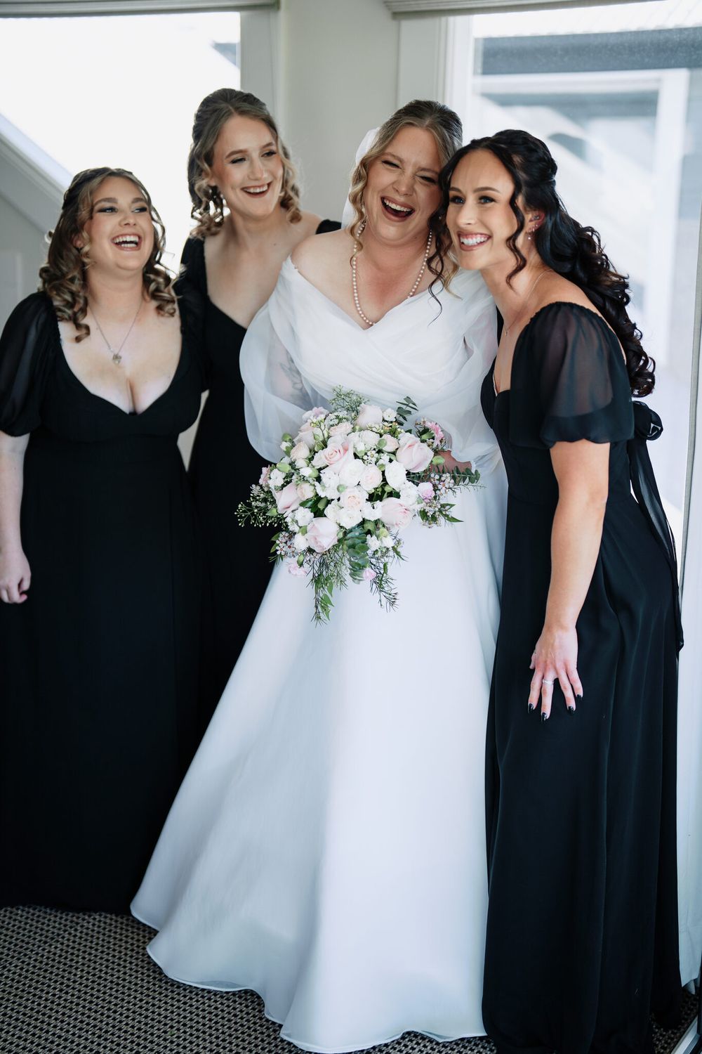 Smiling bride in a white gown holding a pink and white bouquet, laughing with three bridesmaids in black dresses before the wedding.