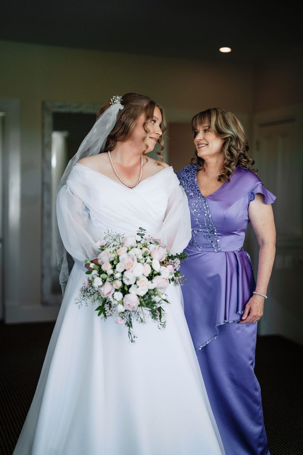 A bride in a white off-the-shoulder gown and veil holds a pink rose bouquet beside a smiling woman in a lavender satin dress.