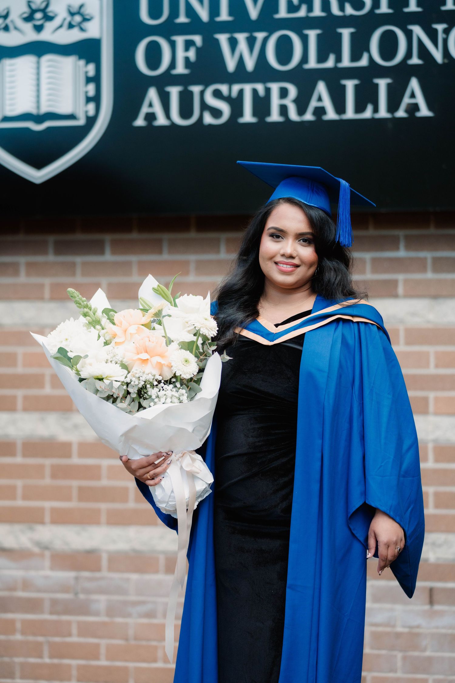 Smiling graduate in a blue gown and cap holding a white and peach bouquet in front of a University of Wollongong Australia sign.