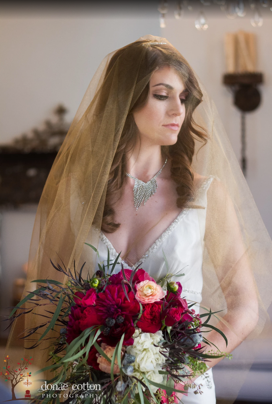 Bride in white gown and veil holding vibrant red and pink bouquet, elegant necklace, soft indoor lighting.