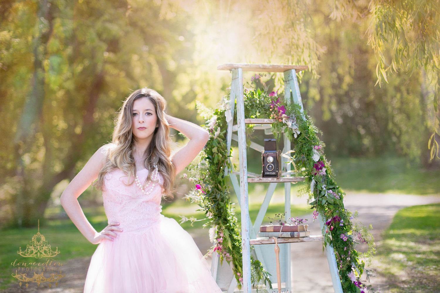 Young woman in pink tulle gown poses outdoors near floral-decorated ladder in golden sunlight.
