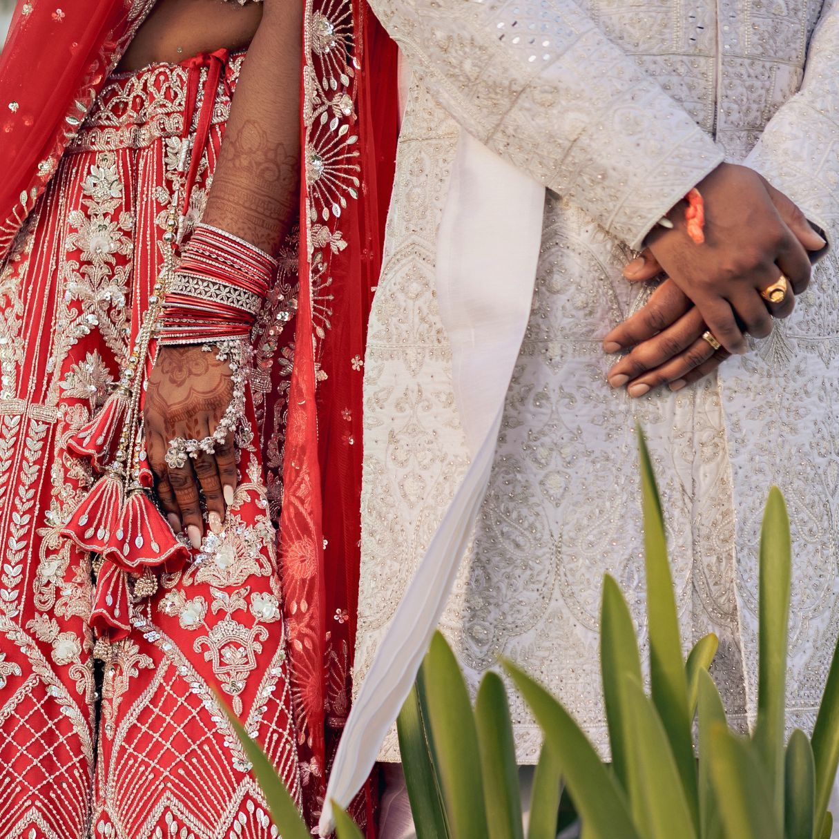 Indian bride in red embroidered lehenga with bangles and groom in white sherwani holding hands at wedding ceremony.