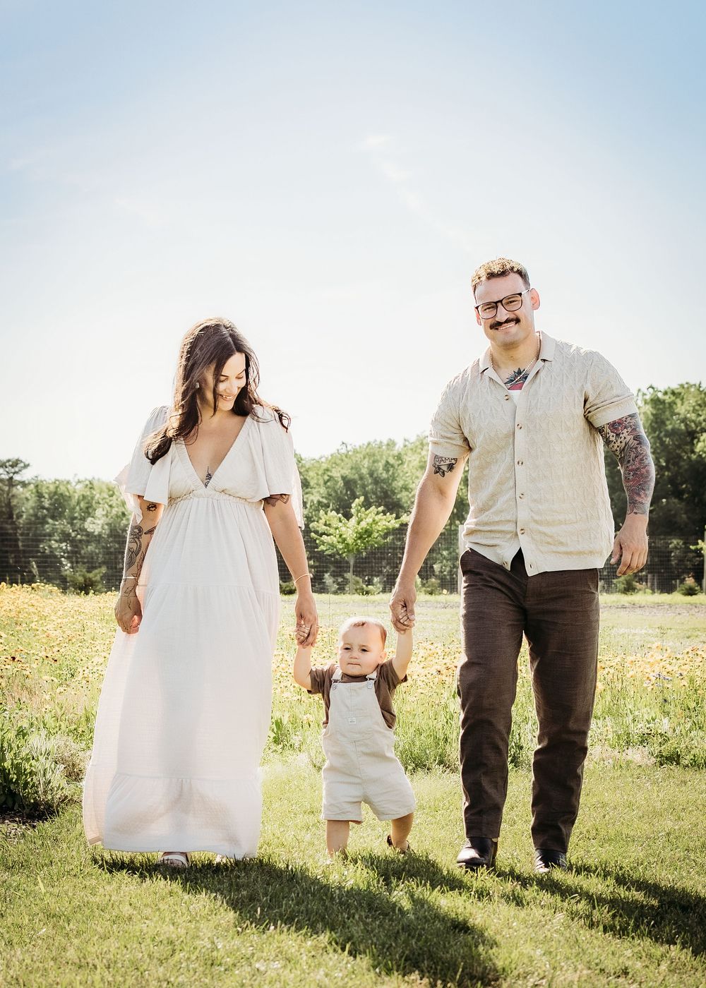 Mom and dad walking through a field while holding their toddler’s hands.