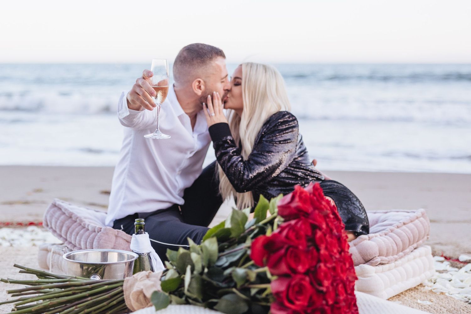 Newley engaged woman kissing her fiancé sitting behind a bouquet of roses as he holds a glass of champagne on the beach