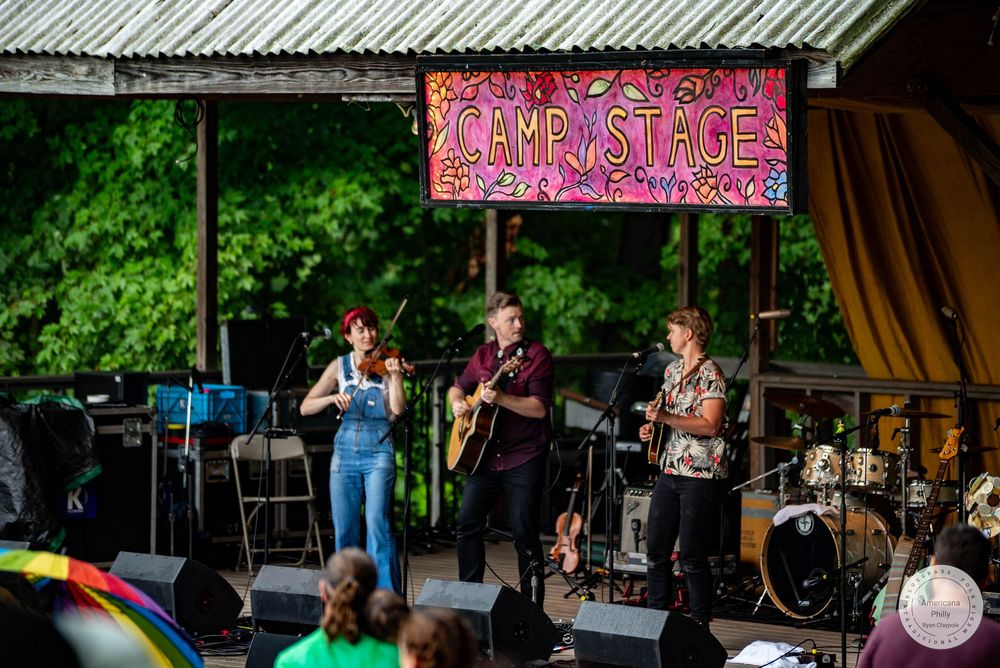 Three musicians perform on the Camp Stage at an outdoor music festival, playing violin, guitar, and mandolin.
