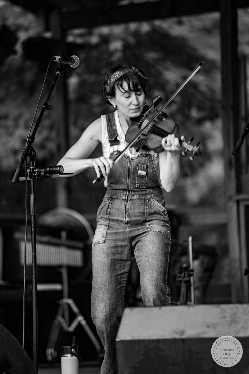 Woman in denim overalls playing violin on outdoor stage, black and white performance photo.