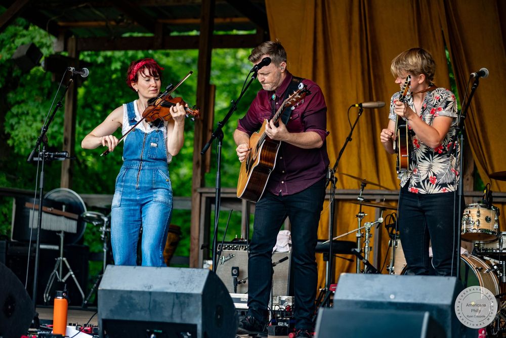 Three musicians perform on outdoor stage, woman playing violin, man with acoustic guitar, and another with microphone.