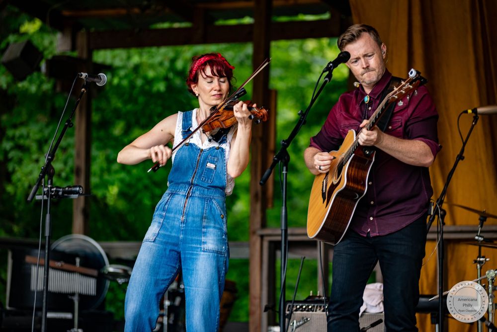 A woman playing violin and a man playing acoustic guitar perform on an outdoor stage at a live music event.
