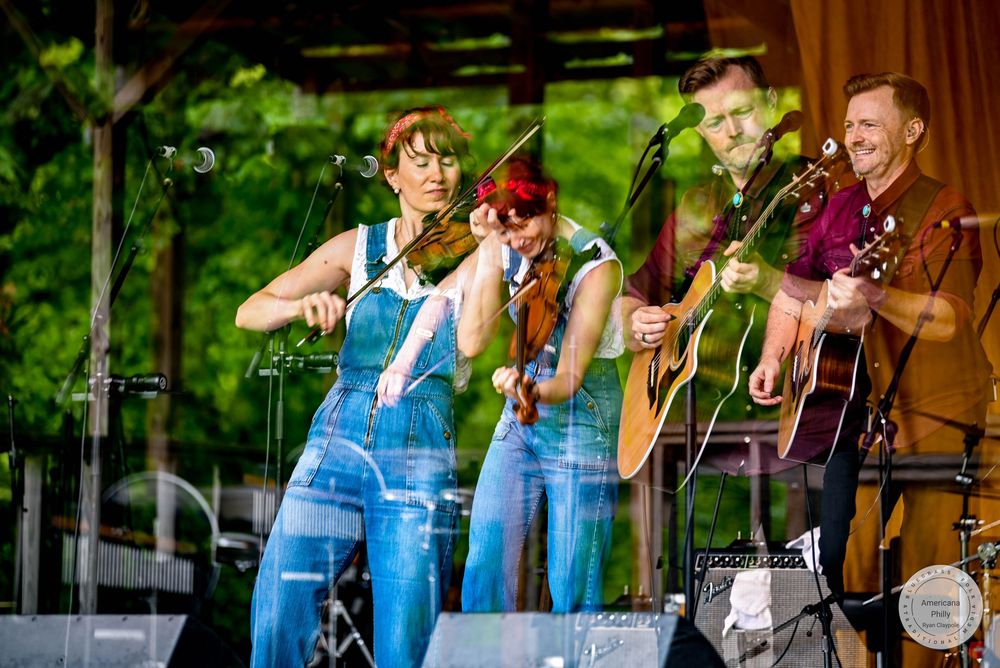 Live bluegrass band performing outdoors, two female violinists and male guitarist on stage at a summer concert.
