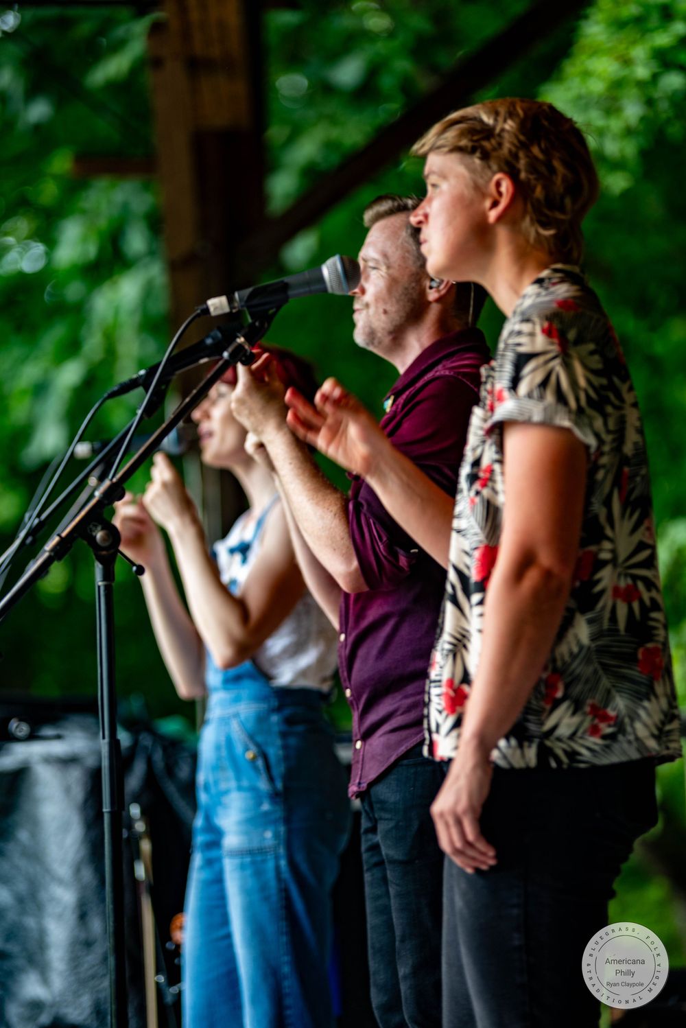 Outdoor concert performers singing at microphones on stage surrounded by lush green trees.
