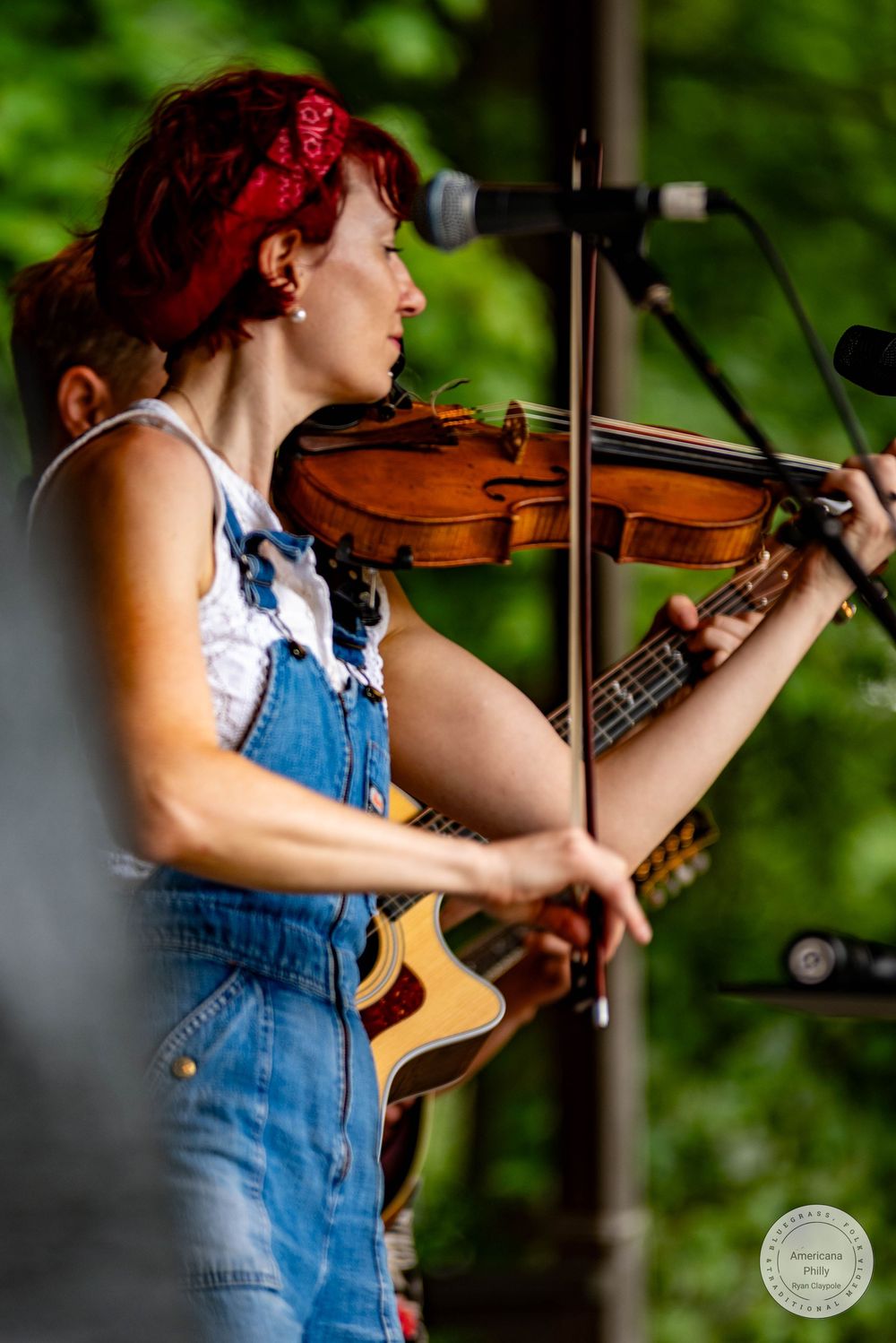 Woman with red hair playing violin on stage, wearing denim overalls at an outdoor music festival.