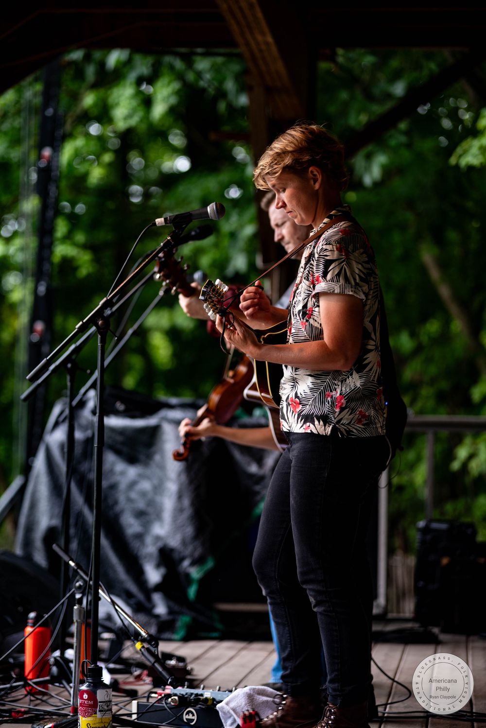 Woman playing acoustic guitar on outdoor stage with microphone, wearing floral shirt, performing at live music event.