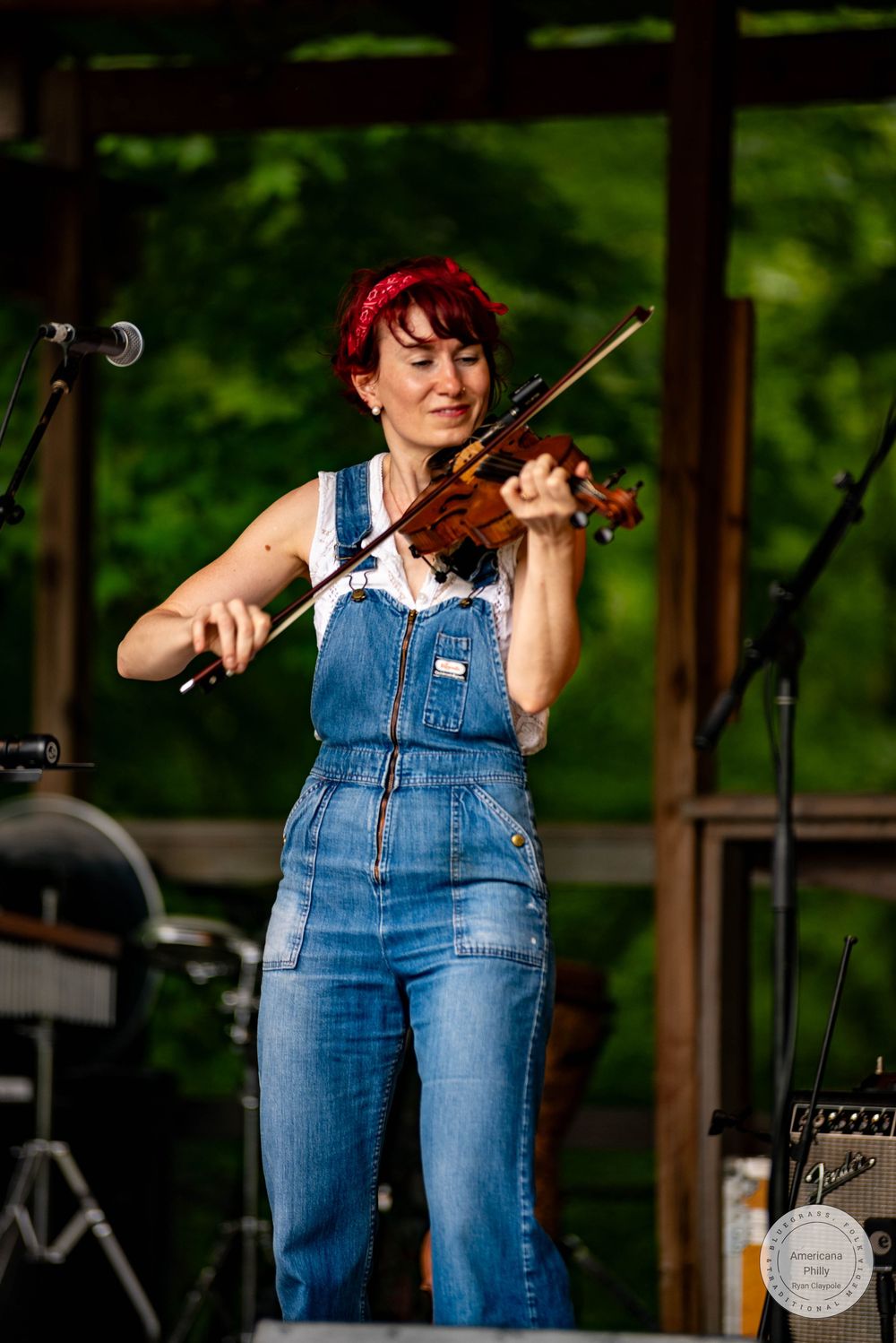 Woman in denim overalls playing violin on outdoor stage with green trees in background.