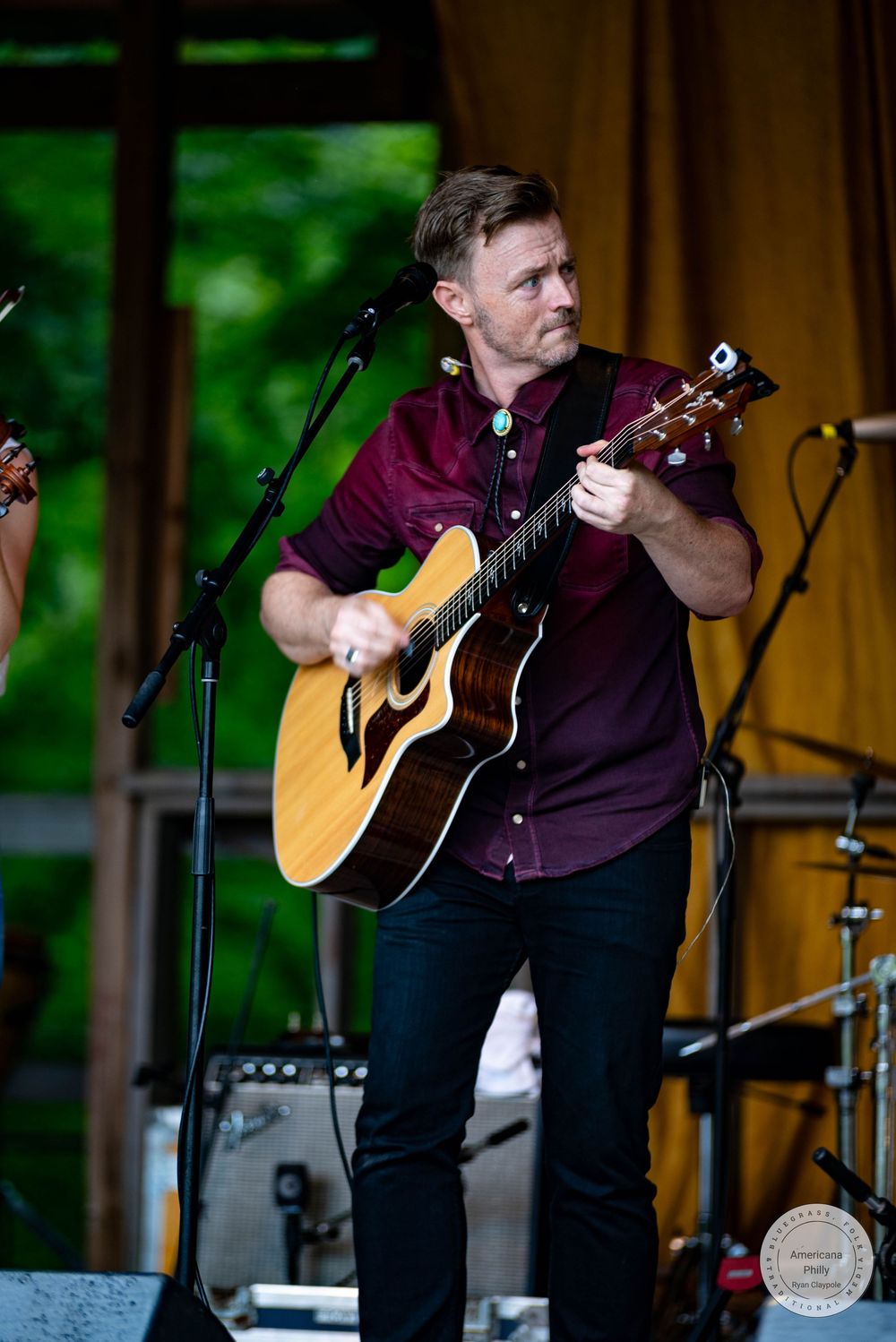 A man in a burgundy shirt plays acoustic guitar on an outdoor stage surrounded by lush green trees.