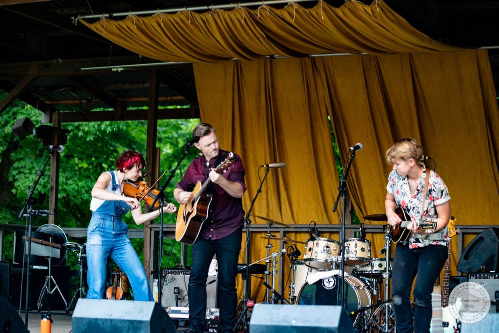 Live outdoor folk band performance with violinist, guitarist, and bassist on stage with yellow curtain backdrop.