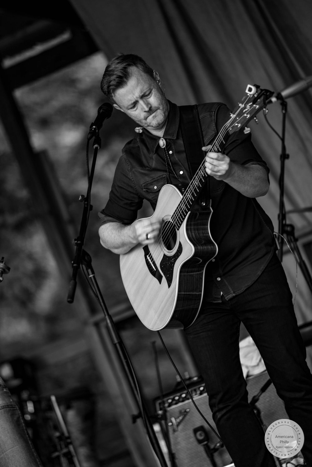 Black and white photo of a man passionately playing acoustic guitar on stage at an outdoor concert.