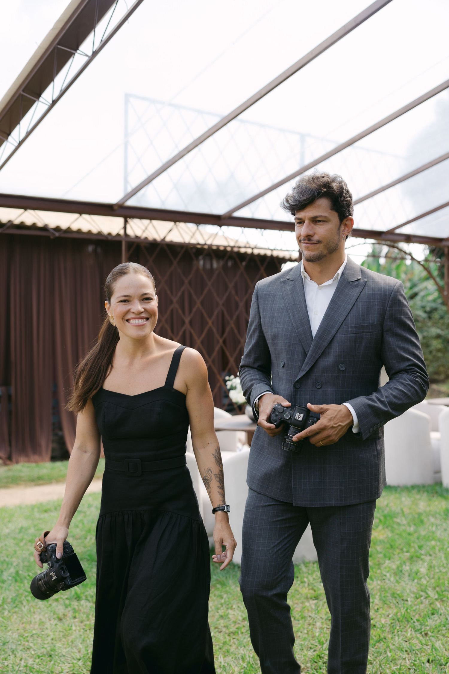 A smiling woman in a black dress and a man in a gray suit walk outdoors, both holding cameras.