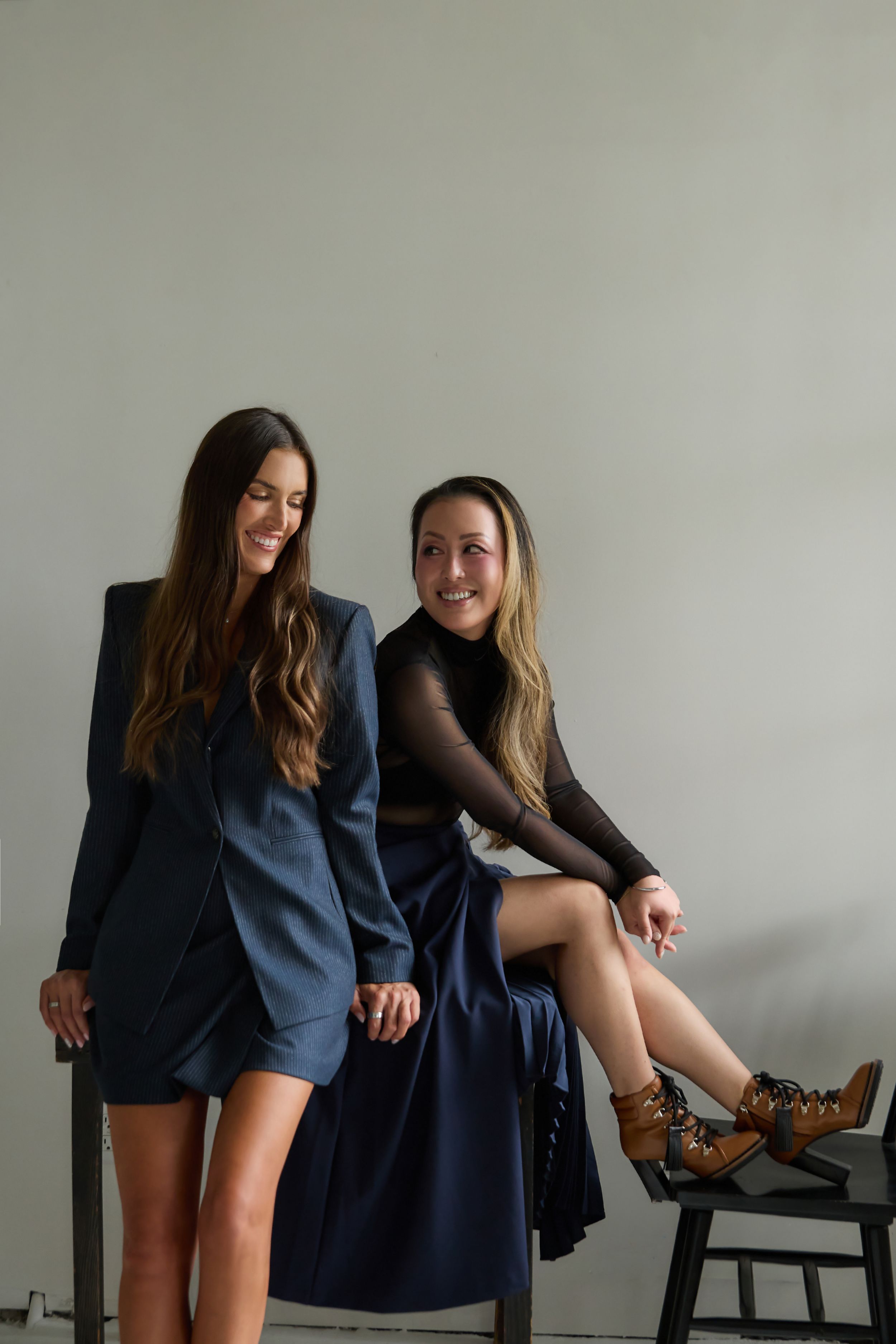 Two stylish women posing together, one in a blue blazer dress and one in a black mesh top with navy skirt and boots.