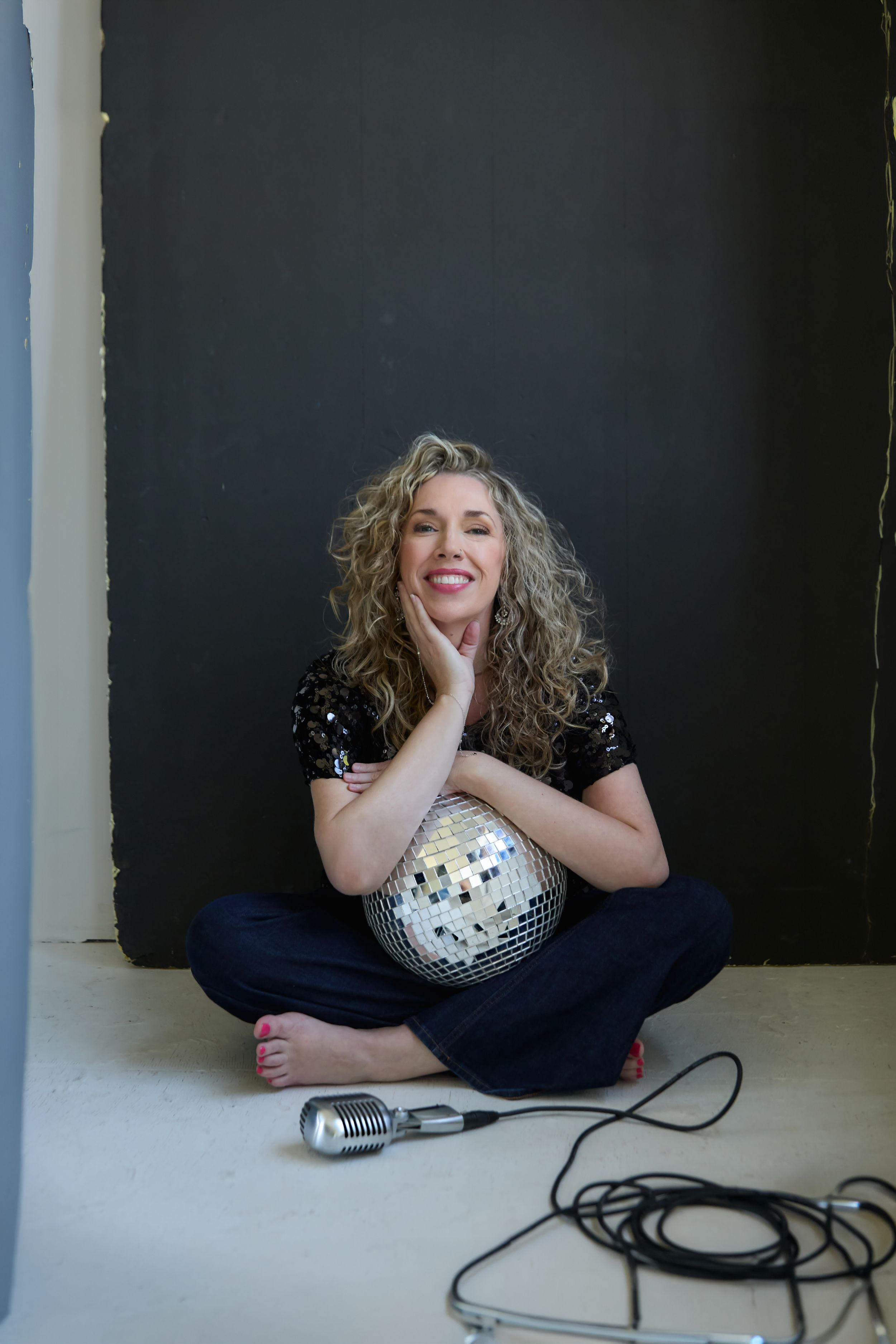 Smiling woman with curly hair sits cross-legged on floor holding disco ball, microphone nearby, against dark wall.