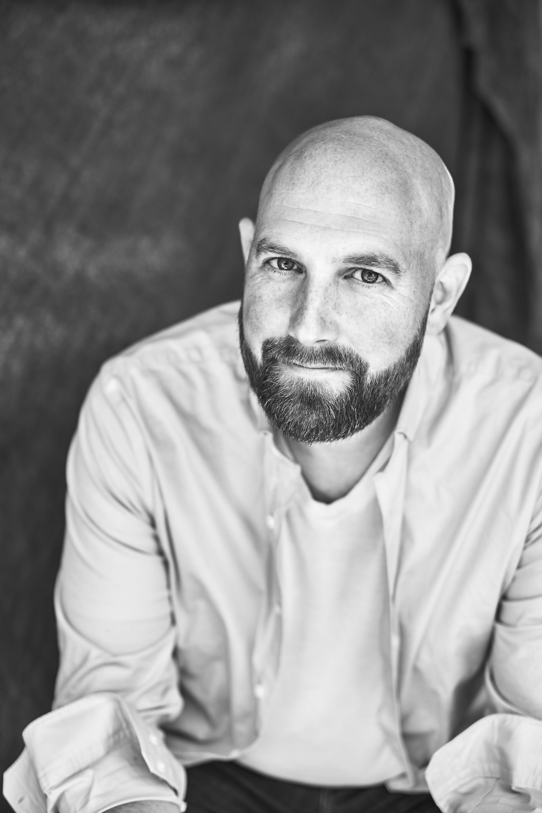 Bald man with beard wearing white shirt poses in black and white portrait photo.