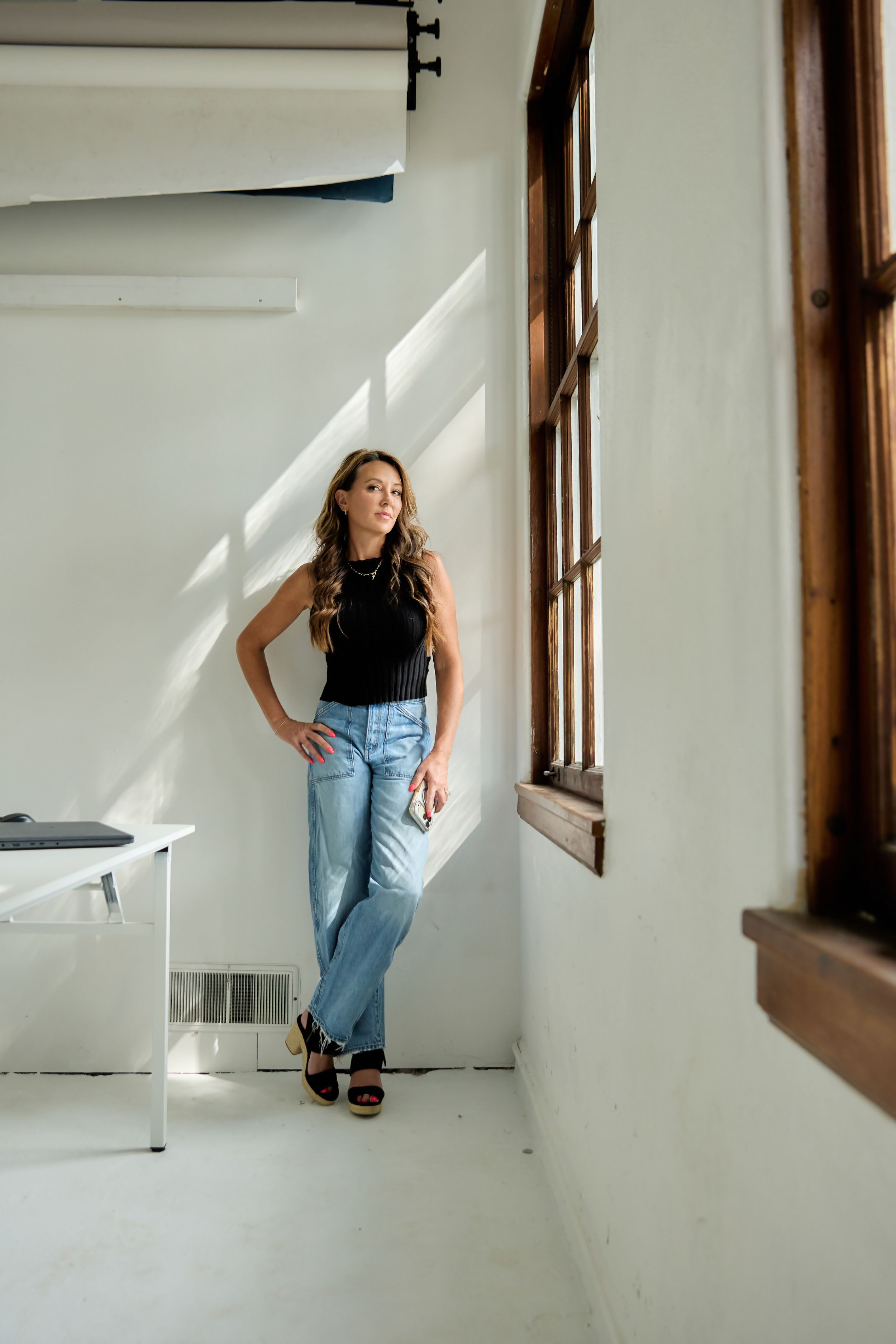 Woman in black sleeveless top and light blue jeans leaning against white wall near wooden windows in bright studio space.