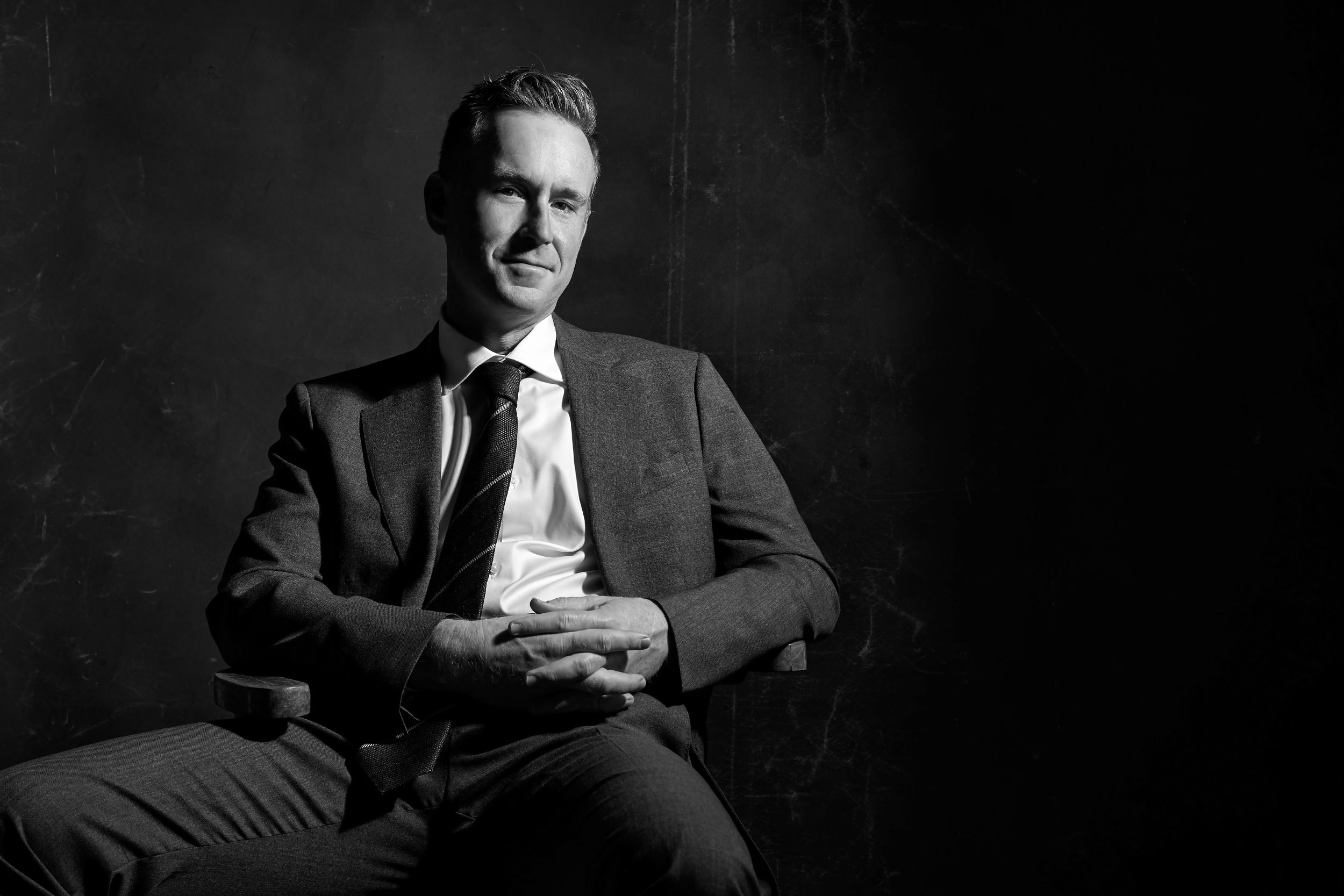 Black and white portrait of a confident businessman in a suit sitting relaxed with hands clasped in a dark studio.