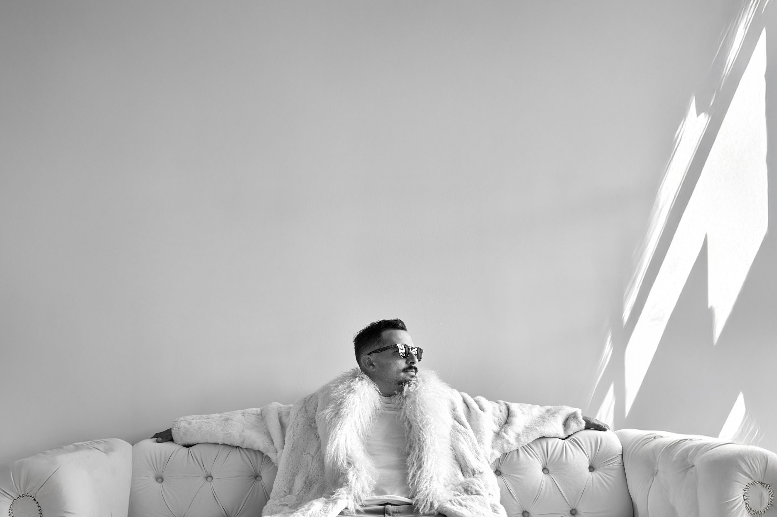 Black and white photo of a person sitting on a tufted sofa wearing sunglasses and a fur coat in a bright room.