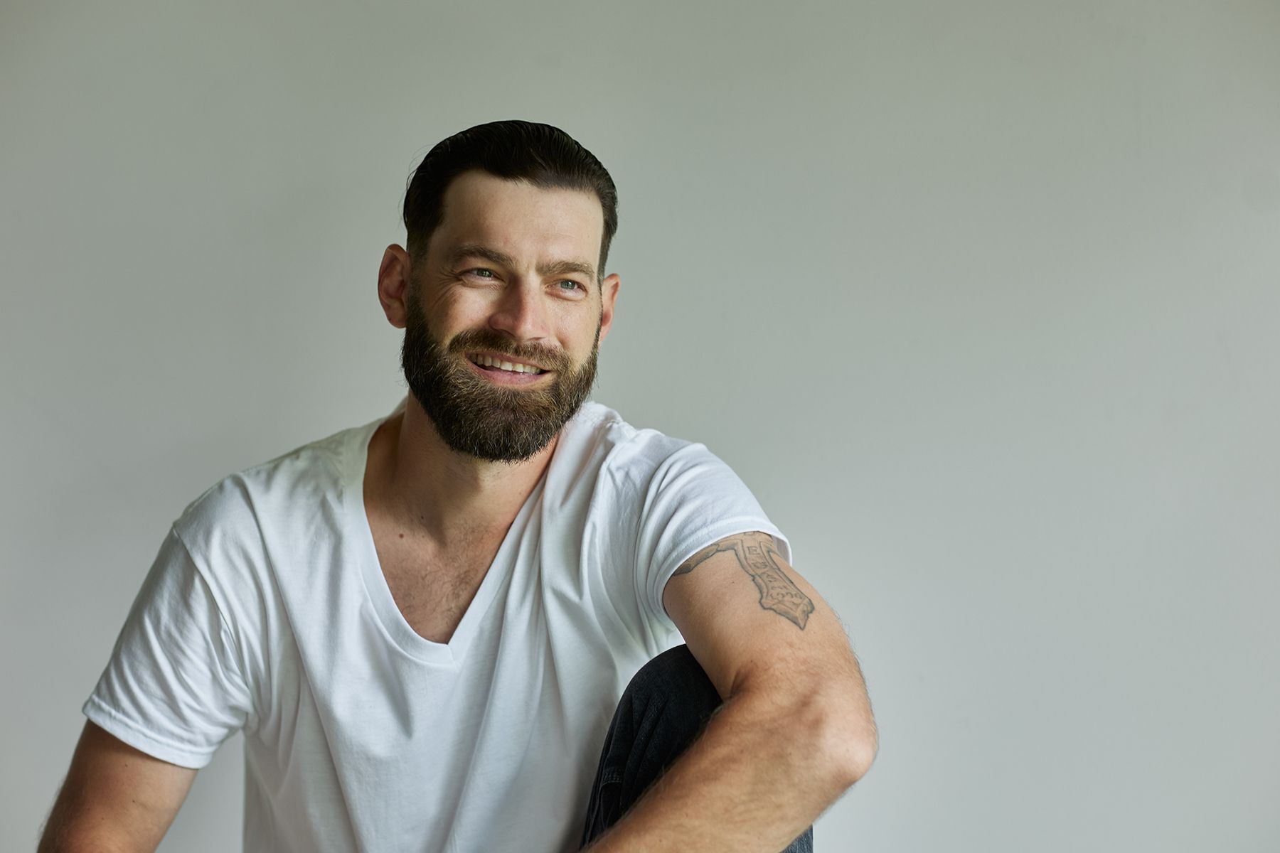 Smiling bearded man in white t-shirt with arm tattoo posing casually against a light gray background.