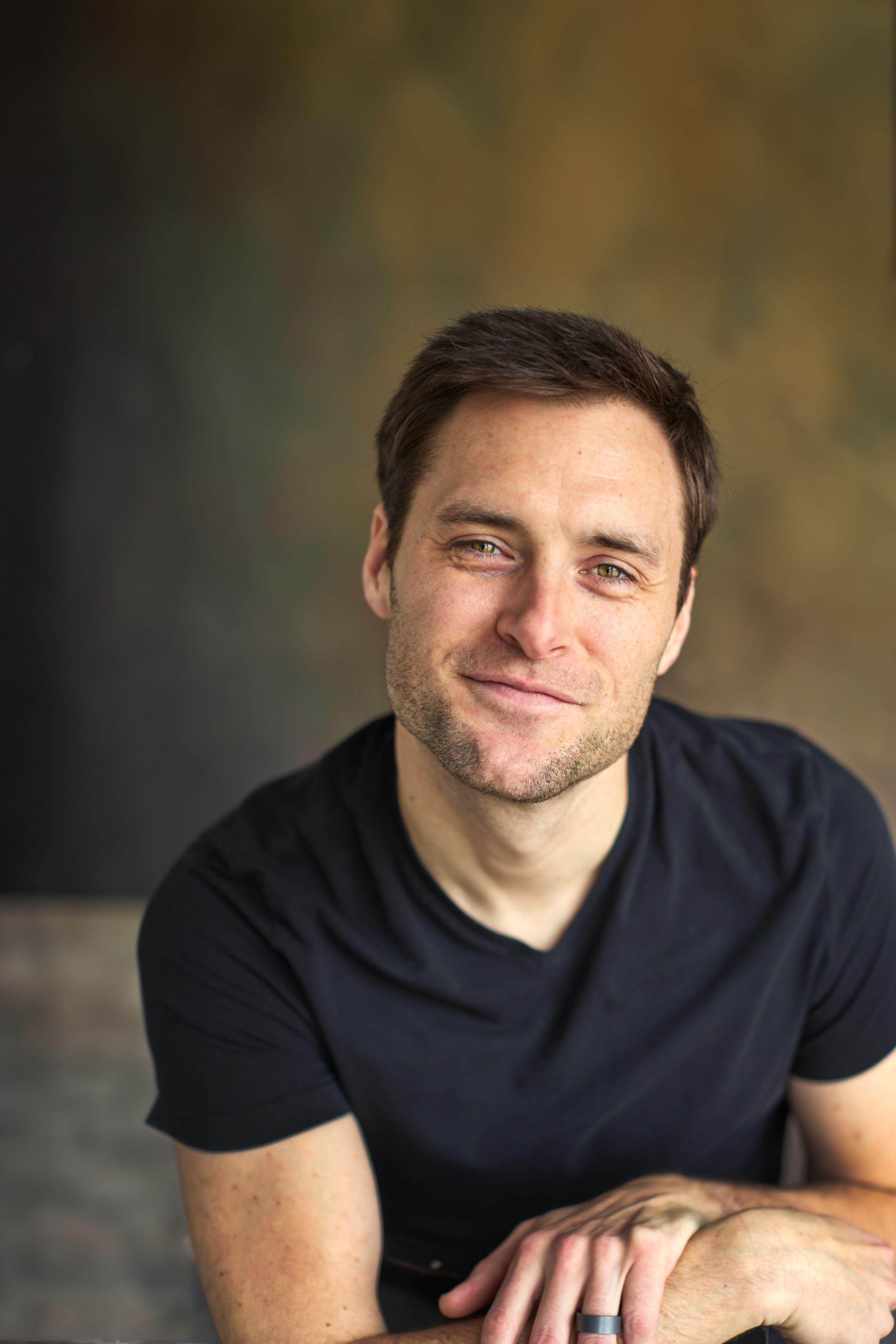 Smiling man in black t-shirt with short brown hair posing casually against a textured dark background.