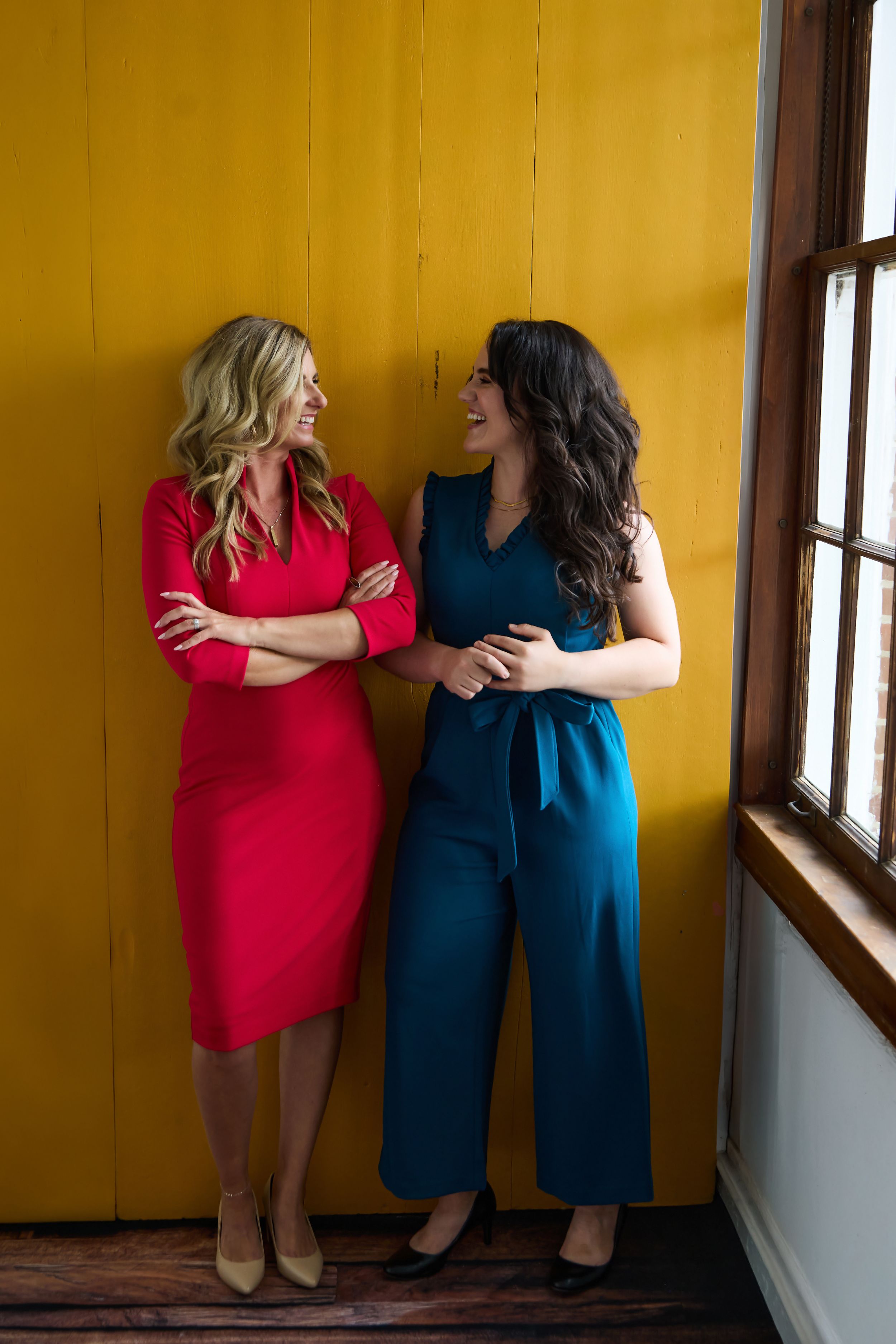 Two women smiling and laughing together, one in a red dress and one in a teal jumpsuit, standing against a yellow wall.