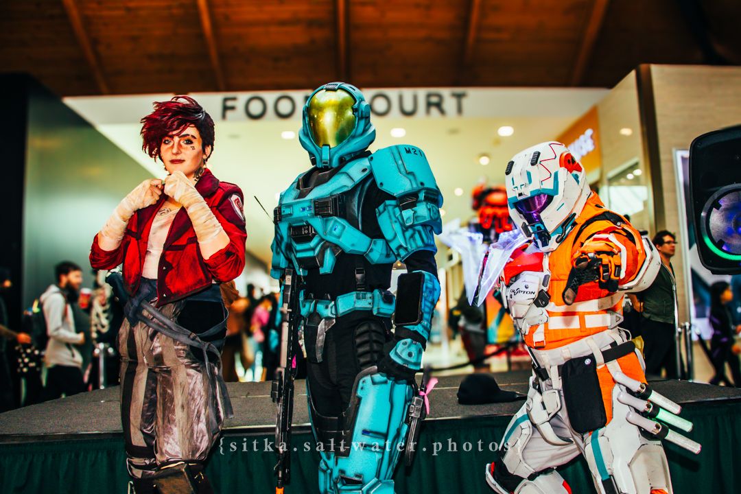 Three cosplayers pose near a food court, featuring armored Halo and Star Wars X-Wing pilot costumes at a convention.